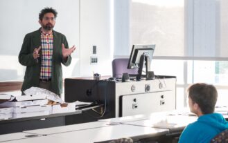 A man stands in front of a whiteboard talking with a computer to his right, while a student sits in front on him.