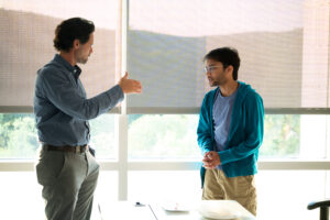 A man speaks with a male student in front of a window. Both are standing.