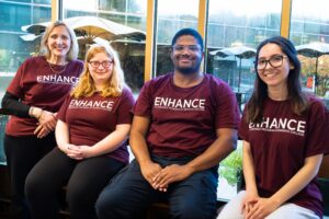 A group of four people with maroon shirts reading ENHANCE sit in front of a window.