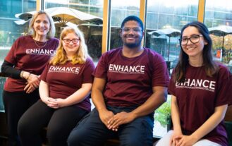 A group of four people with maroon shirts reading ENHANCE sit in front of a window.