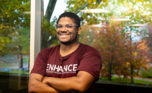 A man wearing a maroon ENHANCE shirt crosses his arms and smiles.