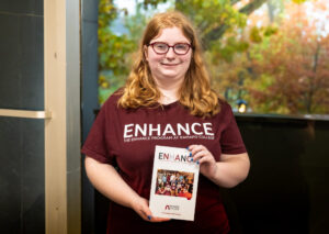 A woman with a maroon shirt that reads ENHANCE holds a brochure for ENHANCE.