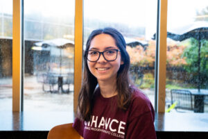 A woman with glasses smiles and sits in front of a window.