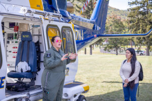 A flight nurse in a dark green jump suit talks in front of a helicopter. A woman looks on.