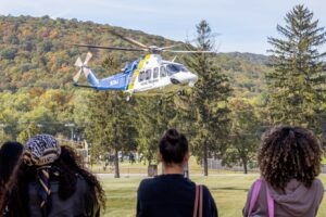 A helicopter hovers in front of a mountain. Three women stand and watch. The backs of their heads are in the picture