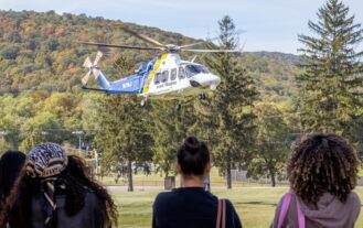 A helicopter hovers in front of a mountain. Three women stand and watch. The backs of their heads are in the picture