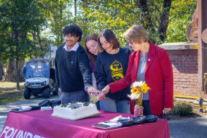Four people stand behind a table with a cake on it. Together they reach to cut the cake.