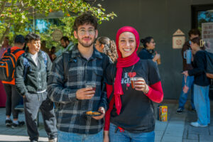 Two college students stand side by side outside with drinks in their hands.