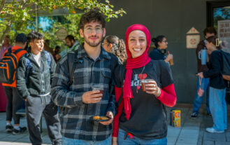 Two college students stand side by side outside with drinks in their hands.