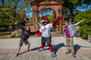 Two people stand in front of an arch outside with Rocky a college mascot standing between them.