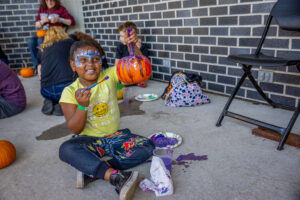 A young girl with face paint on holds up a paint brush and a painted pumpkin and smilles.