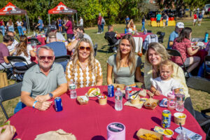 A man, three women, one with a baby on her lap, sit at a table outside with a maroon tablecloth. 