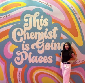 A woman stands in front of a multi-colored wall that reads, This Chemist is Going Places