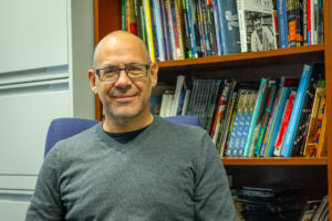 A man in a gray sweater and glasses sits in front of a bookcase filled with books.