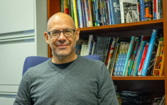 A man in a gray sweater and glasses sits in front of a bookcase filled with books.