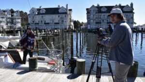 A college student stands with a tri-pod and a video camera in front of water and houses.