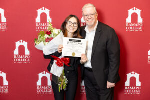 A young woman holding flowers and a certificate smiles next to a man in a suit. They stand in front of a Ramapo College of New Jersey backdrop.