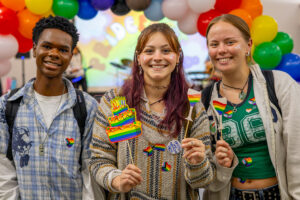 Three students stand side by side, smiling. The girl in the middle holds a rainbow flag. There is a rainbow balloon arch behind them.