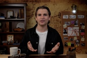 A man sits behind a desk with his hands open. He has a coffee cup on his desk, and a map in the background.