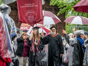 A woman in a cap and gown holds a huge maroon gonfalon and walks.