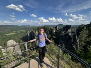 A male college student stands in the corner of guardrails. Behind him is an amazing mountain view and a bridge behind him to the right.