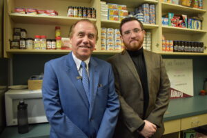 Two men in suits stand shoulder to shoulder smiling. They are in front of shelves of food.