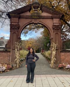 A woman stands outside in front of a large arch with her hand on her hip.