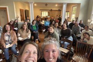 Two women smile in the foreground, with others sitting in chairs behind them in a room.