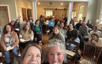 Two women smile in the foreground, with others sitting in chairs behind them in a room.