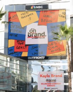 A billboard with red, yellow, blue and white Post-It notes is displayed in front of a glass building.