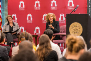 Two women sit on stage on chairs in front of a maroon background with Ramapo College of New Jersey logos. In front of them is an audience. A podium is to the left.