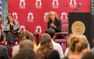 Two women sit on stage on chairs in front of a maroon background with Ramapo College of New Jersey logos. In front of them is an audience. A podium is to the left.