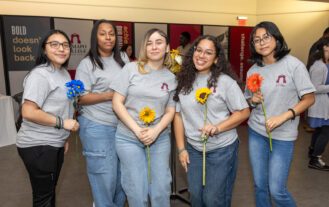 Five young women wearing gray t-shirts and jeans hold flowers and smile. They are standing shoulder to shoulder inside.