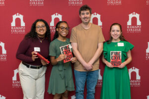 A group of four people pose in front of a maroon wall with Ramapo College logos on it. Three of them hold books.