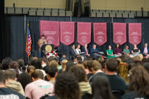 A man stands on stage at a podium, while others sit on stage. A crowd is in front of the  stage.