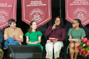 A group of thre people sit on a stage. One holds a microphone and the others listen.