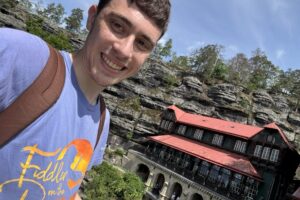 A male college student with a brown backpack smiles for a selfie in front of a historic building next to cliffs.