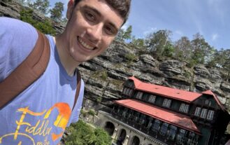 A male college student with a brown backpack smiles for a selfie in front of a historic building next to cliffs.