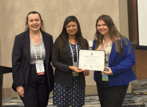 Three women stand side by side. Two hold either end of a certificate.