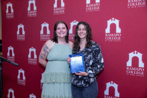 Two women stand in front of a Ramapo College banner. One holds an award.