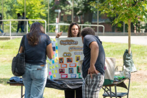 A college student stands behind a table with a poster on it and speaks to two other students in front of her.