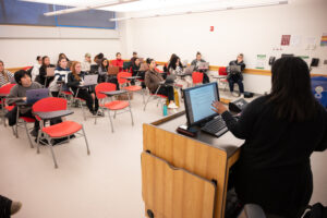 A female professor stands in front of a class. She is at a computer. The class is sittind in front of her in red desks.