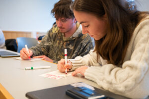 Two college students holding markers sit at a table and color on cards.
