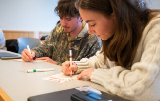 Two college students holding markers sit at a table and color on cards.