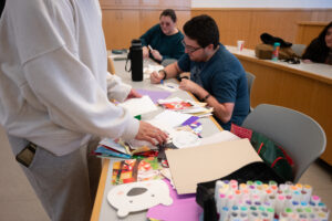 Two college students sit at a table covered in cards and markers, while another stands looking through cards.