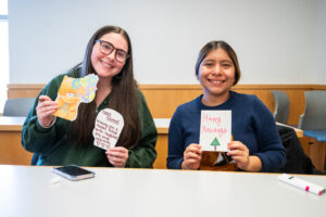Two female college students smile while they sit at a table and hold up holiday cards that they created.
