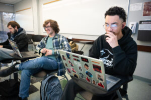 Three students sit in a classroom in front of laptops.