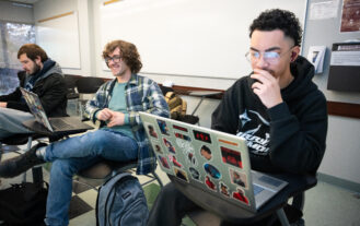 Three students sit in a classroom in front of laptops.