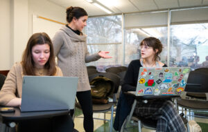 Two students sit down in front of laptops as a teacher speaks to them.