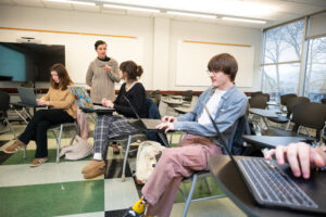 Students sit at desks in front of laptops as a teacher stands and speaks.